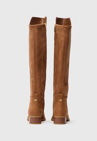 Bottes en suede marron au-dessus du genou avec une finition lisse, des accents en métal doré et un talon empilé. Détail de fermeture éclair à l'arrière.