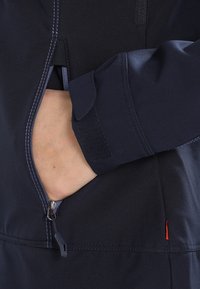 Marinblå jacka med en dragkedjeficka, mjukt material och kontrasterande sömmar. Inkluderar justerbara ärmslut med kardborre.