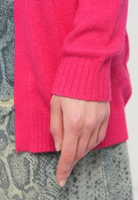 Mano rilassata al fianco, indossando un maglione lavorato a maglia di un vivace rosa brillante con polsini a coste e pantaloni grigio-blu a fantasia con una toppa strappata.