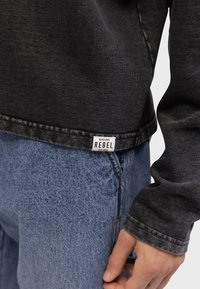Chemise à manches longues en denim noir avec un dégradé de couleurs, poignets cousus. Portée sur un jean en denim bleu clair avec une étiquette de marque visible.