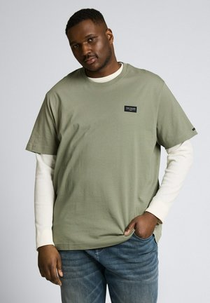 Mann trägt ein khakigrünes Kurzarmhemd über einem weißen Langarmhemd und blaue Jeans, steht mit einer Hand in der Tasche.