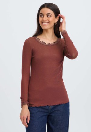 Femme souriante, portant un haut marron à manches longues avec bordure en dentelle au col et aux poignets, associé à un jean bleu, sur un fond uni.