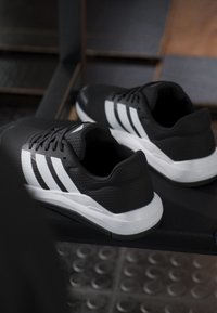 Chaussures de sport noires avec semelles blanches et trois bandes blanches latérales, posées sur un banc de gym sombre dans un environnement d'entraînement.