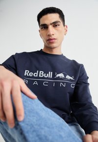 Jūrinio stiliaus džemperis su ilgomis rankovėmis ir apvaliu kaklu, ant kurio yra baltas „Red Bull Racing“ logotipas ir du įsikandę buliukai.