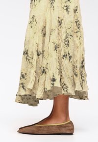 Zapato plano de ante marrón con punta afilada y ribetes en contraste de color verde lima, combinado con una falda amarilla con patrones florales en negro y plisados.