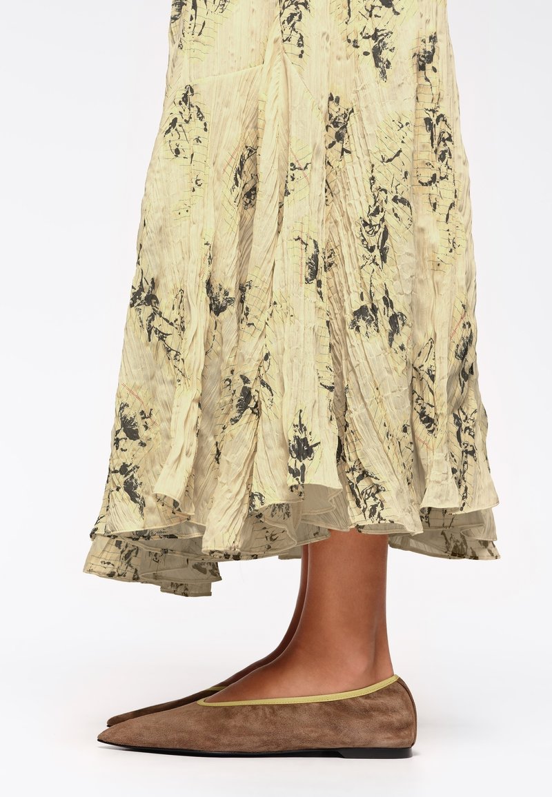 Zapato plano de ante marrón con punta afilada y ribetes en contraste de color verde lima, combinado con una falda amarilla con patrones florales en negro y plisados.