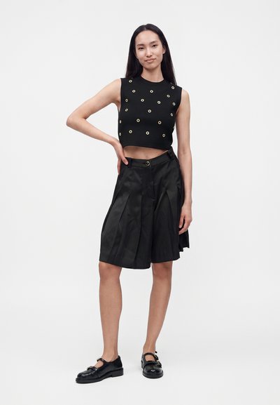 Jeune femme portant un crop-top noir sans manches avec des œillets circulaires et un short noir plissé taille haute jusqu’aux genoux avec des chaussures noires.