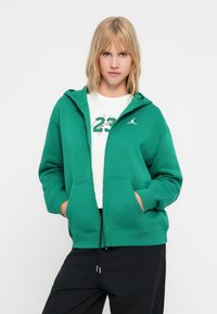 Sudadera verde con cremallera delantera, de tela suave, con dos bolsillos laterales y logo blanco. Combinada con una camiseta gráfica blanca y pantalones negros.