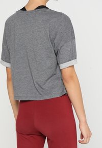 Persona con una camiseta corta gris de manga corta y puños enrollados, y pantalones rojos de cintura alta, de pie frente a un fondo blanco liso.