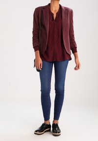 Femme portant un blazer et une chemise bordeaux, un jean skinny bleu, des chaussures plateforme noires, et tenant un sac à bandoulière noir à chaîne.