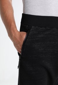 Schwarze Jogginghosen mit strukturierter Oberfläche, ausgestattet mit einem gerippten elastischen Bund und Reißverschlusstaschen. Dargestellt mit einer Hand in der Tasche.