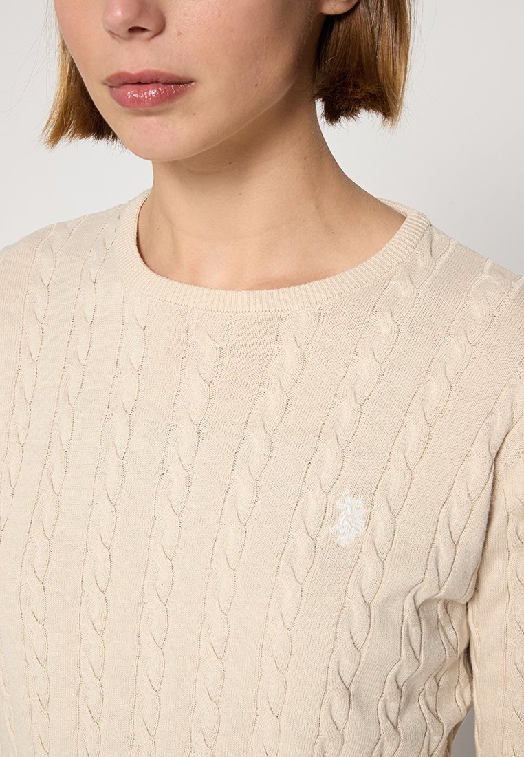 Frau in einem beigen, grob gestrickten Pullover mit Rundhalsausschnitt und einem kleinen, weißen, gestickten Logo auf der Brust.