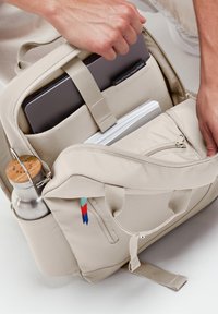 Zaino beige con più tasche, una manica per laptop e un portabottiglie. Presenta accenti di colore contrastante sulle zip.