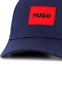 Casquette bleu marine avec une visière courbée, comportant un patch rectangulaire rouge bien visible portant l'inscription "HUGO" en noir, fabriqué en tissu texturé.