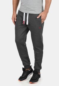 Herrengraue Jogginghosen aus weichem Material, mit schmalem Schnitt, elastischem Taillenbund mit weißen Kordeln und kleinem roten Logo.