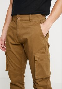 Hnědé cargo kalhoty se střihem na rovné nohavice, s několika kapsami a zapínáním na knoflíky. Vyrobené z odolné látky s hladkým, texturovaným povrchem.