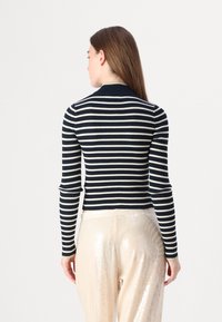 Woman facing away wearing a navy and white striped fitted long-sleeve top and shiny beige sequined pants against a plain background.