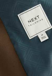 Zwei Stoffmuster: eines in Dunkelbraun, das andere in Dunkeltürkis mit einem geometrischen Muster. Ein weißes Etikett trägt die Aufschrift "NEXT TAILORING" und die Größe M.