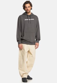 Gray hoodie with a front pocket and white logo, paired with light-colored, loose-fitting pants and black footwear. Neutral background.