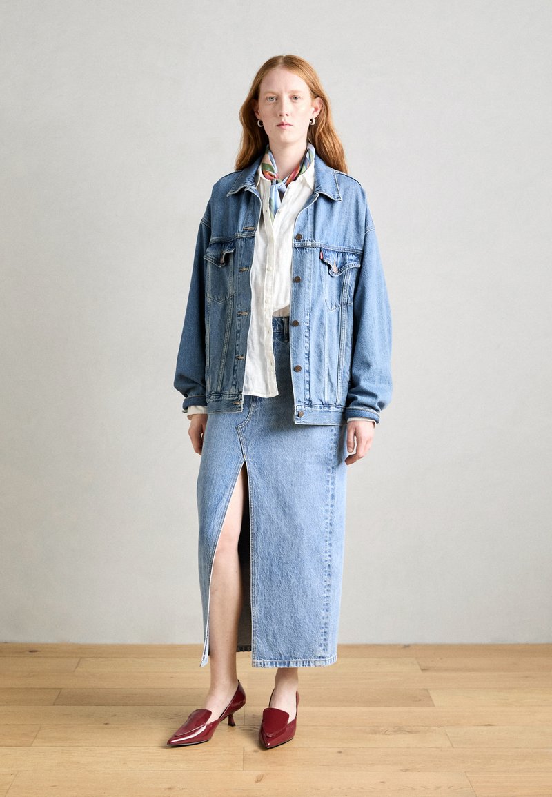 Ensemble en jean composé d'une veste et d'une jupe bleu clair, avec une fente haute sur la jupe. Chaussures rouges et chemise blanche en dessous. Conçu pour être superposé.