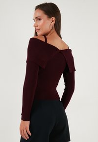 Sieviete ar gariem taisniem matiem, ģērbusies tumši burgundijas krāsas ribotā svārka bez pleciem un melnās biksēs, raugās sānis ar lielajām aļģēm ausīs.