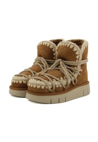 Stivali alla caviglia in suede marrone con superficie texturizzata, cuciture decorative in crema e lacci, e suola in gomma grigia chiara.