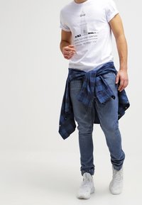 Bílý tričko s grafikou majáku, v kombinaci s modrými džíny a modrou kostkovanou košilí uvázanou kolem pasu. Bílá sneakers dotvářejí outfity.