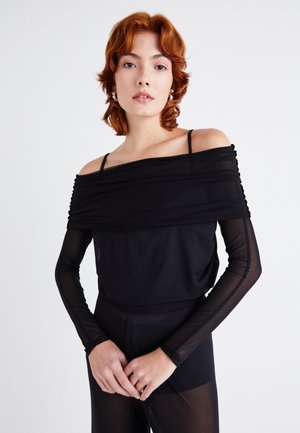 Black off-the-shoulder top with sheer long sleeves, featuring textured fabric and a draped design. Model poses with hands clasped.