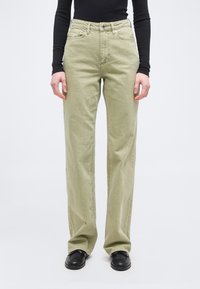 Άτομο που φοράει ανοιχτό πράσινο τζιν με ίσια γραμμή, μαύρο μακρυμάνικο τοπ και μαύρα παπούτσια, στέκεται μπροστά σε λευκό φόντο.