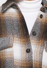 Detailní záběr na hnědou, černou a bílou kostkovanou vlněnou bundu s černými knoflíky na bílém roláku.