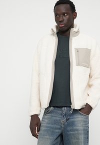 Veste en polaire crème avec un col haut, fermeture éclair à l'avant, et une poche poitrine beige avec une fermeture éclair. Portée sur une chemise vert foncé et un jean en denim.