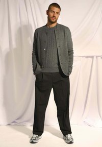 Šedý pletený cardigan přes odpovídající svetr s copánkovým vzorem, spojený s volnými černými kalhotami a šedými sportovními botami s černými akcenty.