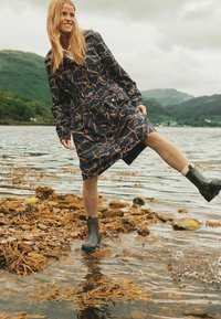 Granatowy płaszcz przeciwdeszczowy w złoty geometryczny wzór, w zestawieniu z beżowymi legginsami i czarnymi gumowymi kaloszami, noszony przez osobę nad brzegiem wody.