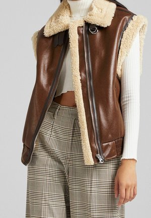 Brown sleeveless shearling vest over white ribbed turtleneck crop top and beige plaid high-waist pants on standing person.