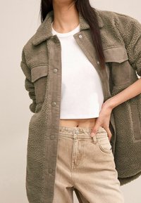 Olijfgroene sherpa jack met corduroy accenten, knoopsluitingen en twee voortassen, gecombineerd met een witte crop top en lichte denim jeans.