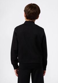 Niño de pie de espaldas, vistiendo un sudadera negra de manga larga y pantalones negros a juego, contra un fondo blanco liso.