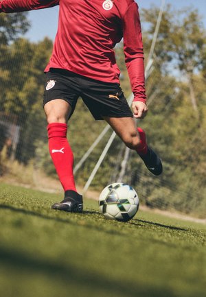 Maglia rossa a maniche lunghe con logo, pantaloni corti neri, calze rosse e scarpe da calcio nere. Giocatore pronto a calciare un pallone da calcio a righe bianche e nere su un prato.