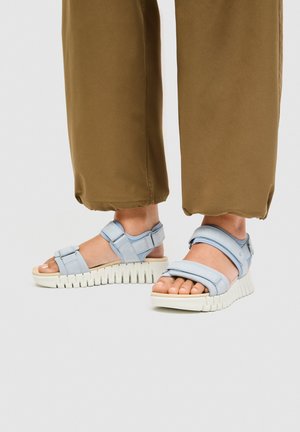 Fødder iført lyseblå sandaler med justerbar rem og tykke hvide rillede såler, kombineret med brune, beskårne bukser med vide ben på en lys baggrund.