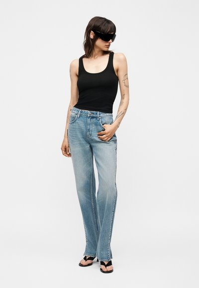 Γυναίκα που φοράει μαύρο αμάνικο τοπ, ανοιχτό μπλε τζιν με τρουκς στο πλάι, μαύρα σανδάλια και μεγάλα μαύρα γυαλιά ηλίου, στέκεται με το χέρι στην τσέπη.