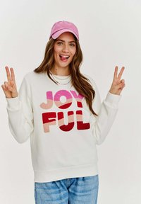 Biely sveter s veľkým, farebným nápisom "RADOSTNÝ" v odtieňoch ružovej a červenej. Nosený s svetloružovou čapicou a džínsami.