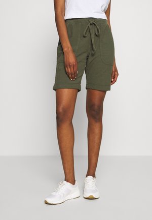 Shorts en coton vert olive avec une taille élastique et un cordon de serrage, comportant des poches latérales et des revers. Assortis avec des baskets blanches.