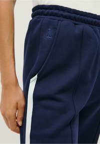 Tumši zilie sporta bikses ar elastīgu jostasvītru, sānu kabatām un baltu joslu gar kabatu šuvēm, ar mazu logotipa detaļu.