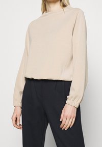 Beżowa bluzka z długim rękawem, wysokim dekoltem i elastycznymi mankietami, wykonana z miękkiego materiału. W połączeniu z czarnymi spodniami o wysokim stanie.