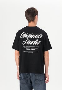 Black cotton t-shirt featuring white text on the back. Design includes "Originals Studio" and additional details in various fonts.