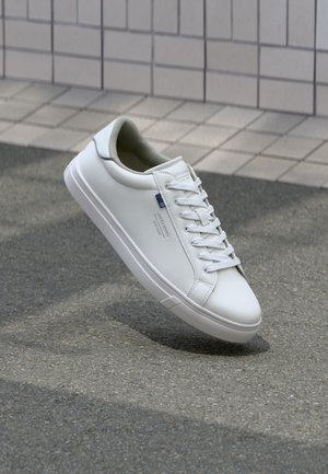 Vita lädersneaker med en slät finish, rund tå, platta snören och en texturerad gummisula; har en liten blå logotyp på sidan.