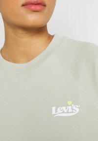 Sweatshirt vert clair avec texture côtelée, arborant un logo Levi's blanc avec un petit cœur jaune en accent sur la poitrine.