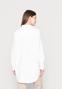 Camisa branca de manga longa com uma bainha ligeiramente curvada, detalhes em botão nos punhos e um corte descontraído, feita de um tecido texturizado.