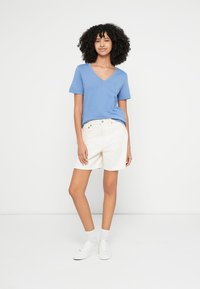 Camiseta de cuello en V azul, pantalones cortos blancos y zapatillas blancas. La camiseta tiene un ajuste holgado, los pantalones cortos llegan hasta la rodilla con un diseño clásico.