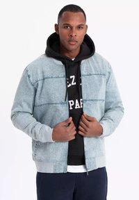 Light blue denim bomber jacket with ribbed cuffs and hem, worn over a black hoodie with white text, featuring a zip-front closure.