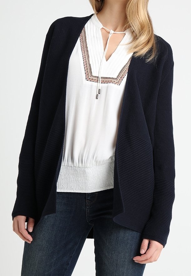 Granatowy prążkowany kardigan noszony na białej bluzce z plisami i ozdobnym przeszyciem, zestawiony z ciemnymi jeansami. Miękka faktura i otwarty przód.